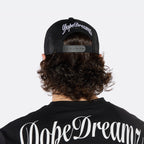 Close-up of a male model wearing the DopeDreamz Interlude Noir black trucker cap featuring a premium white embroidered DDZ monogram, paired with the matching black T-shirt featuring a tonal black embroidered DDZ monogram.
