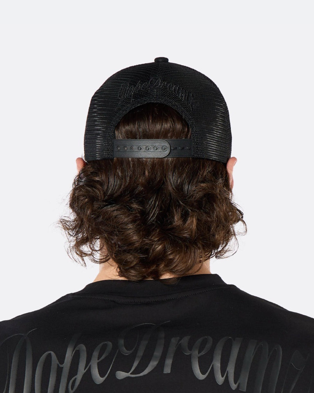 

Rear view of a male model wearing the DopeDreamz Interlude Noir black trucker cap featuring tonal black embroidered DopeDreamz script across the rear mesh panels and an adjustable snapback closure, paired with the matching black T-shirt featuring oversized tonal black DopeDreamz script across the upper back.