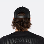 

Rear view of a male model wearing the DopeDreamz Interlude Noir black trucker cap featuring tonal black embroidered DopeDreamz script across the rear mesh panels and an adjustable snapback closure, paired with the matching black T-shirt featuring oversized tonal black DopeDreamz script across the upper back.