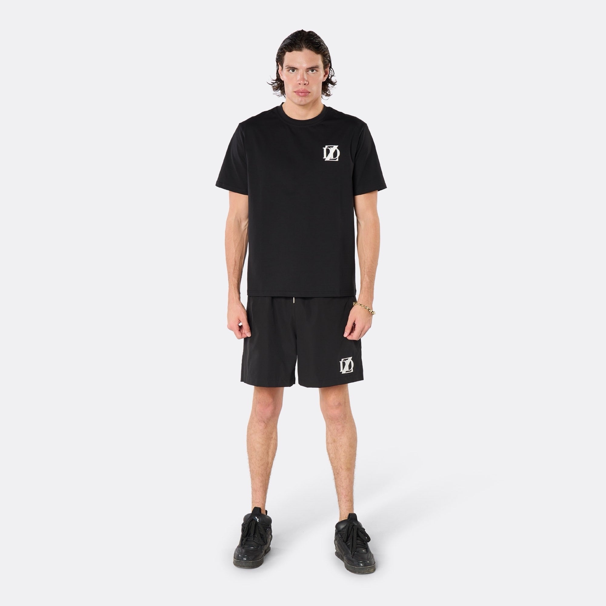 Male model wearing the DopeDreamz Interlude Noir black premium T-shirt with white DDZ chest logo and black shorts with white DDZ logo, front view.