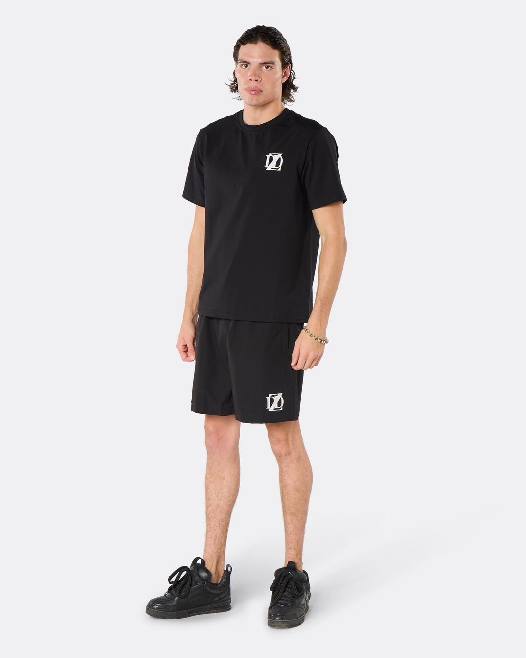 Male model wearing the DopeDreamz Interlude Noir black premium T-shirt with white DDZ chest logo and black shorts with white DDZ logo, three-quarter front view.