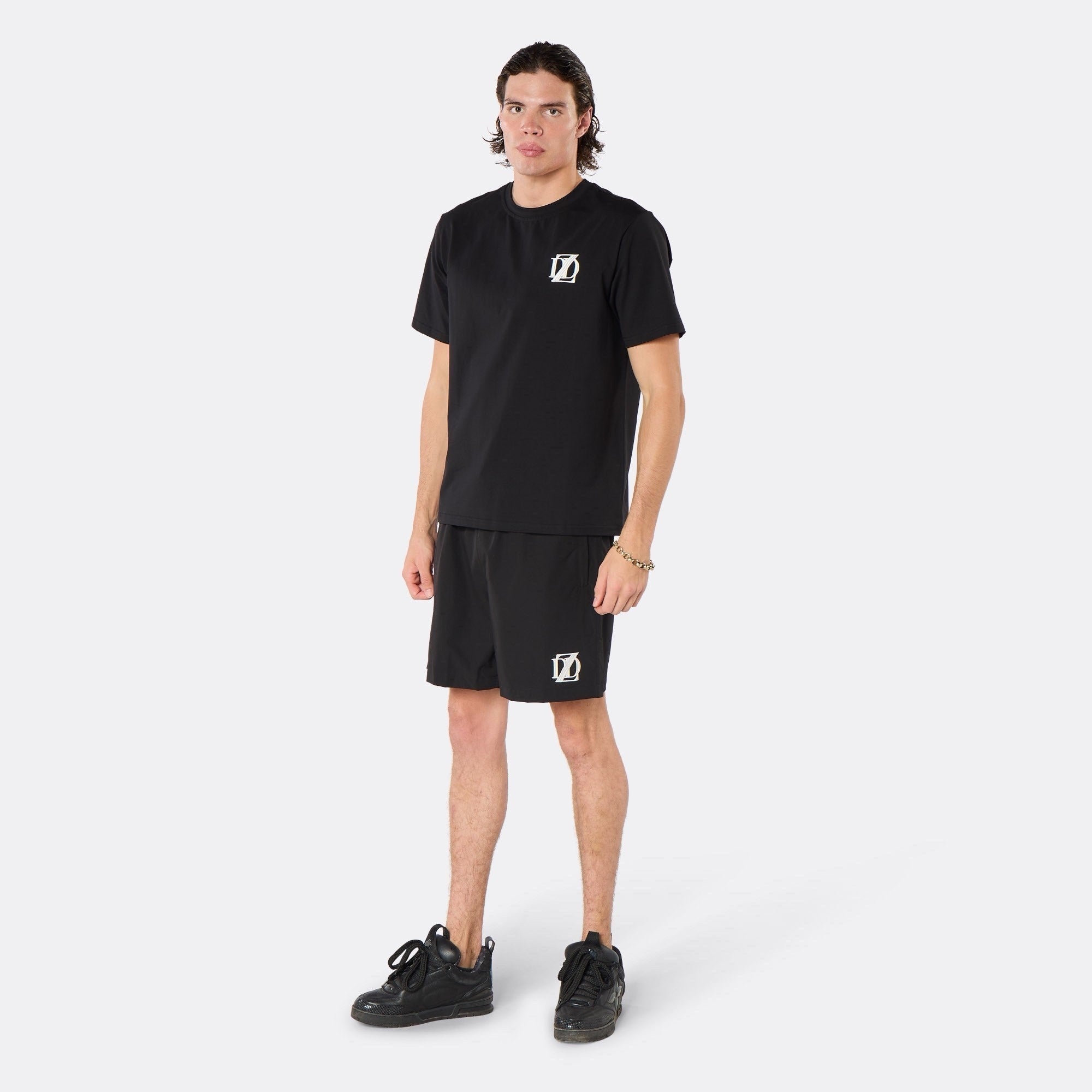 Male model wearing the DopeDreamz Interlude Noir black premium T-shirt with white DDZ chest logo and black shorts with white DDZ logo, three-quarter front view.