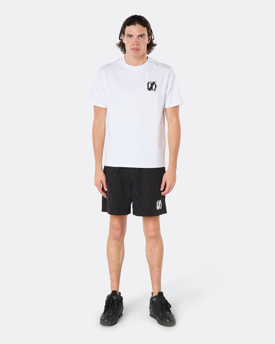 Male model wearing the DopeDreamz white Interlude Noir T-Shirt with black DDZ chest logo and black shorts, with white DDZ logo front view. Paragraph