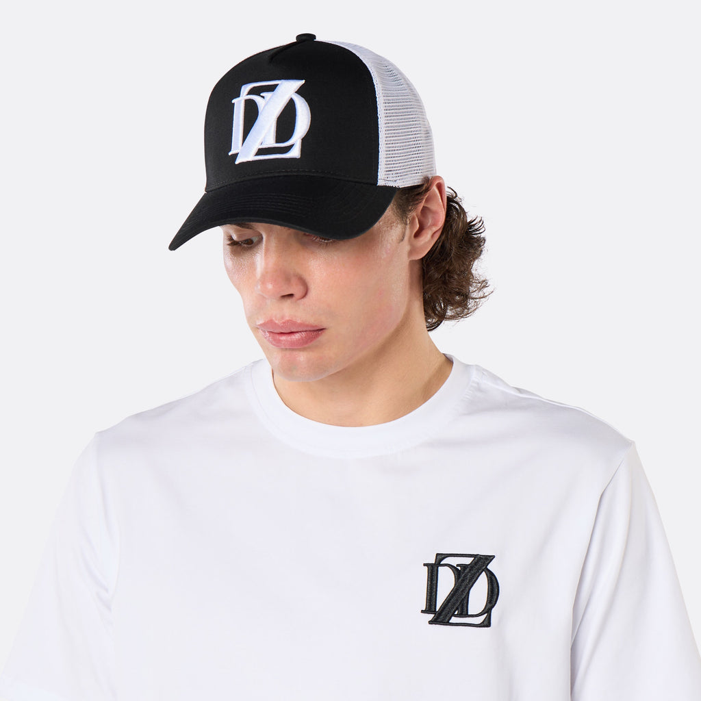 Three-quarter front view of a male model wearing the DopeDreamz Interlude Noir black and white trucker cap with white embroidered DDZ logo and white premium T-shirt with black embroidered DDZ chest logo.