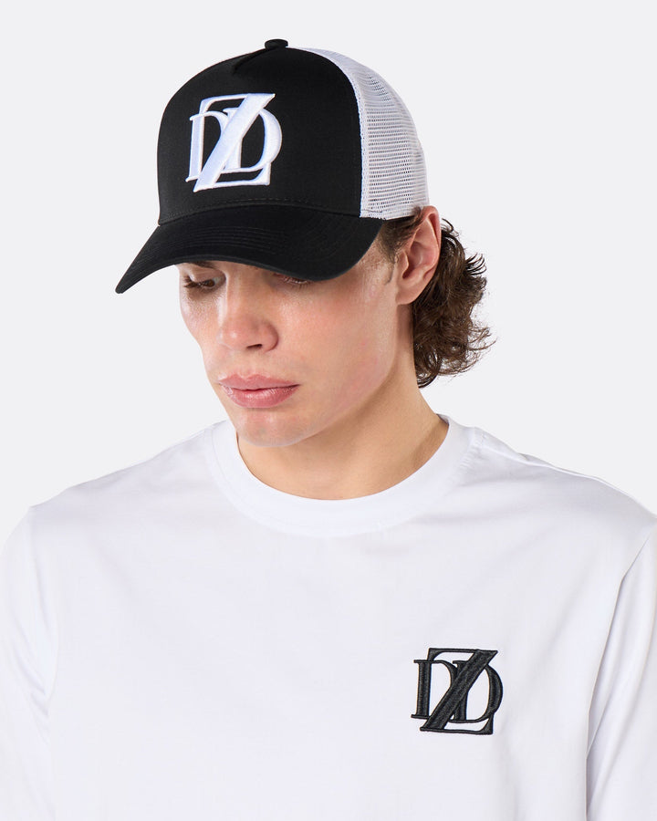 Three-quarter front view of a male model wearing the DopeDreamz Interlude Noir black and white trucker cap with white embroidered DDZ logo and white premium T-shirt with black embroidered DDZ chest logo.