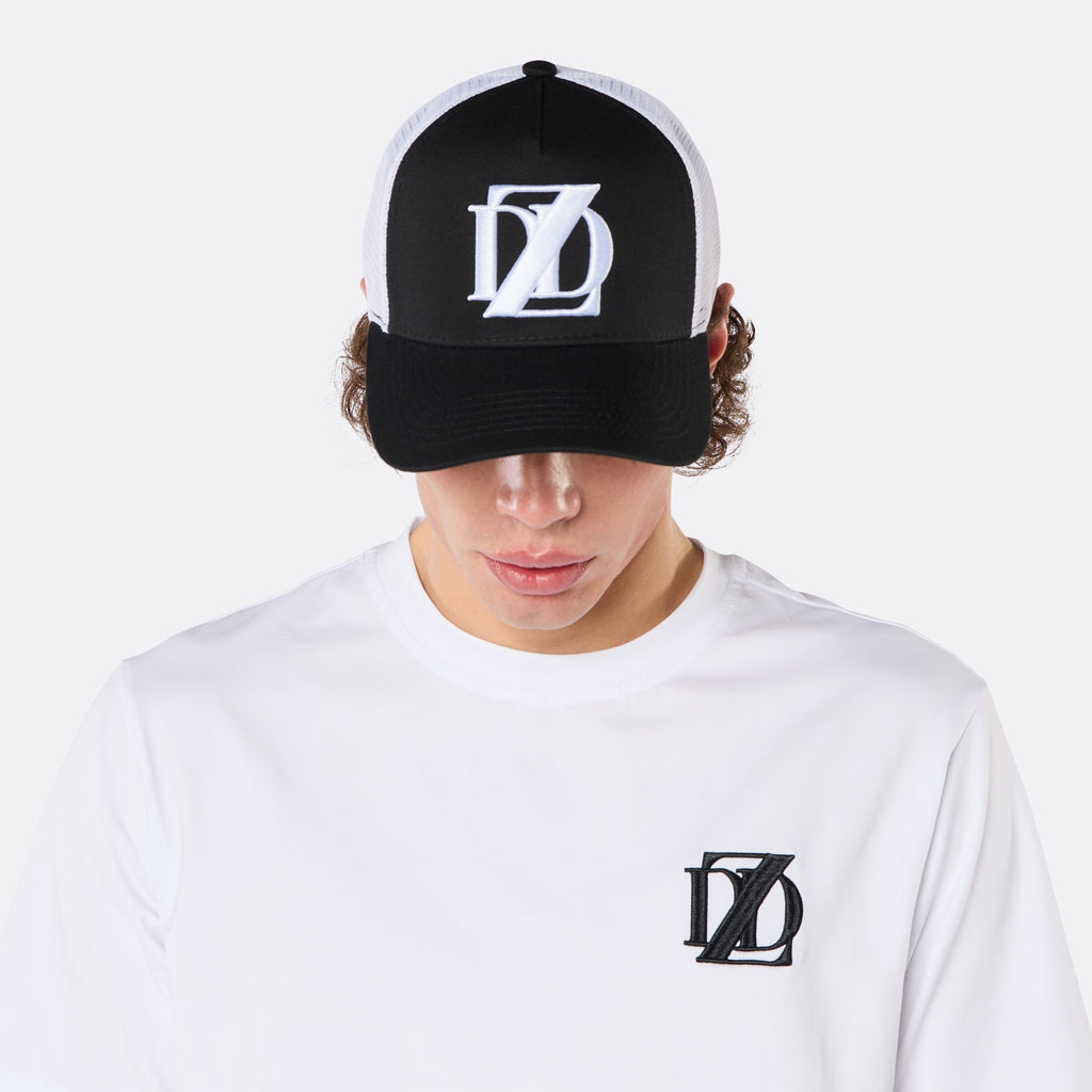 Close-up front view of a male model wearing the DopeDreamz Interlude Noir black and white trucker cap with white embroidered DDZ logo and the white premium T-shirt with black embroidered DDZ chest logo.