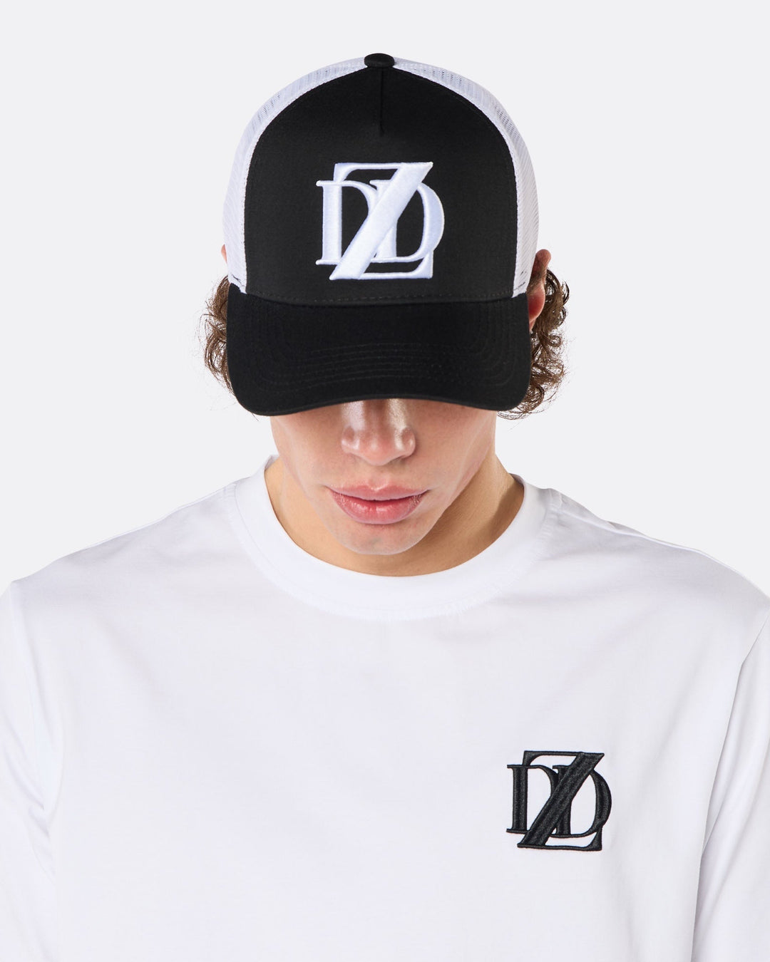 Close-up front view of a male model wearing the DopeDreamz Interlude Noir black and white trucker cap with white embroidered DDZ logo and the white premium T-shirt with black embroidered DDZ chest logo.