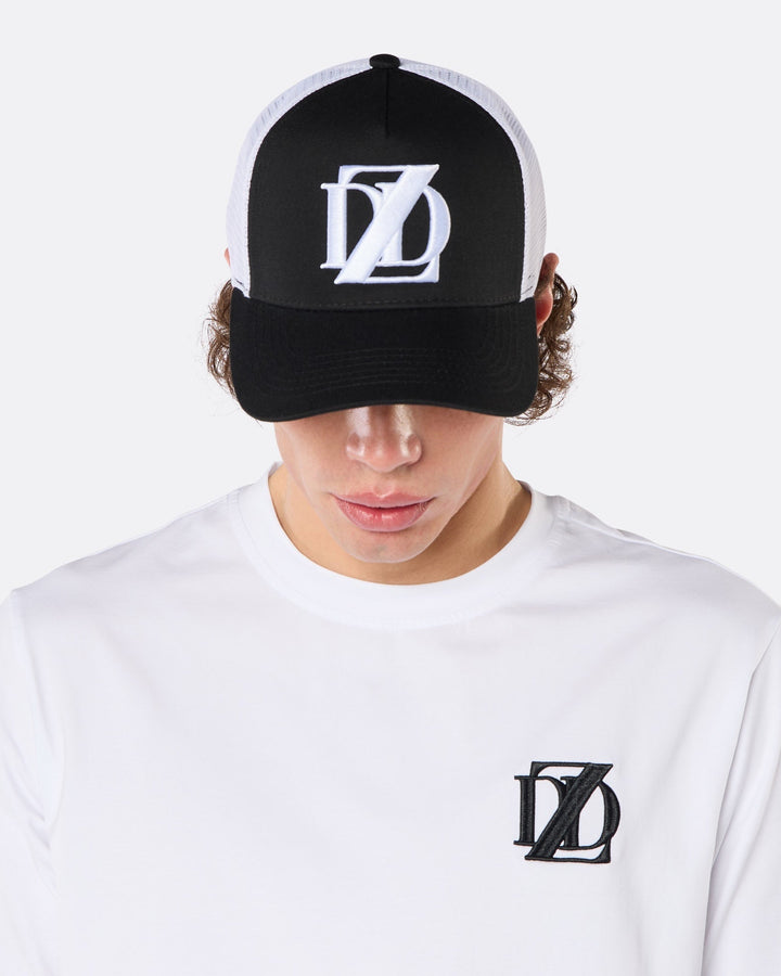 Close-up front view of a male model wearing the DopeDreamz Interlude Noir black and white trucker cap with white embroidered DDZ logo and the white premium T-shirt with black embroidered DDZ chest logo.