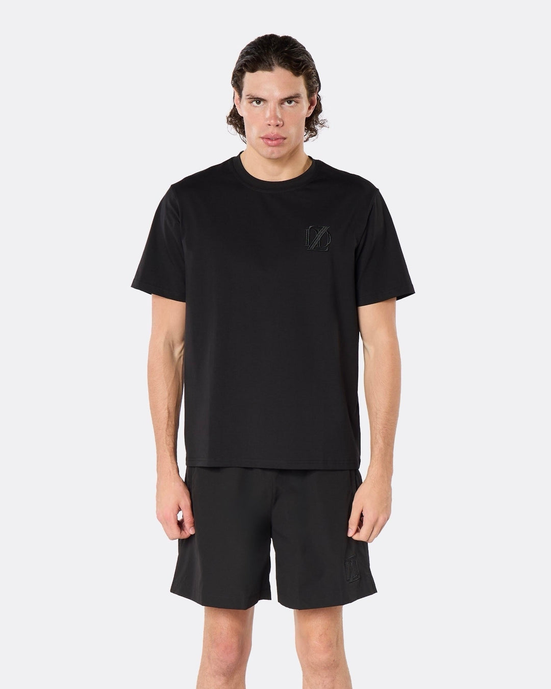 Male model wearing the DopeDreamz Interlude Noir black premium T-shirt with tonal black DDZ chest logo and black shorts with tonal black DDZ logo, close-up front view.
