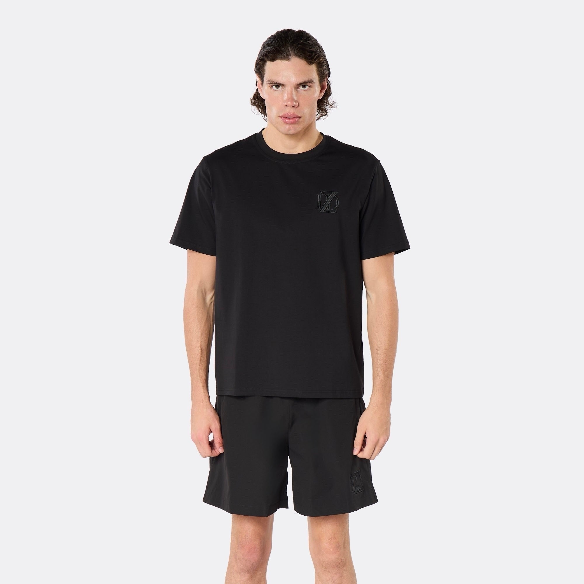 Male model wearing the DopeDreamz Interlude Noir black premium T-shirt with tonal black DDZ chest logo and black shorts with tonal black DDZ logo, close-up front view.