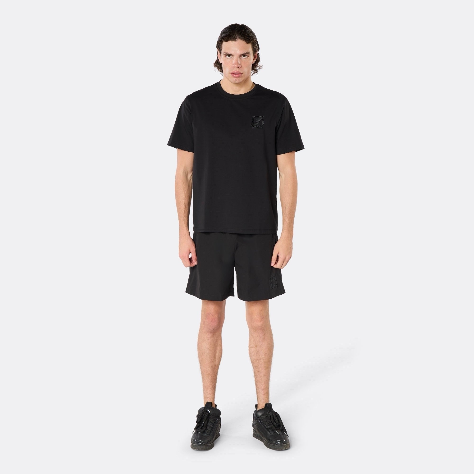 Male model wearing the DopeDreamz Interlude Noir black premium T-shirt with tonal black DDZ chest logo and black shorts with tonal black DDZ logo, front view.