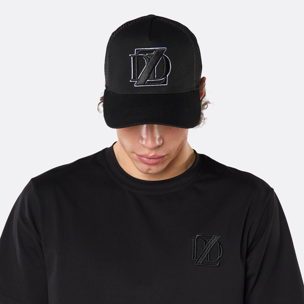 Close-up front view of a male model wearing the DopeDreamz Interlude Noir black trucker cap with tonal black embroidered DDZ monogram, paired with the matching black T-shirt featuring a tonal black embroidered DDZ monogram.
