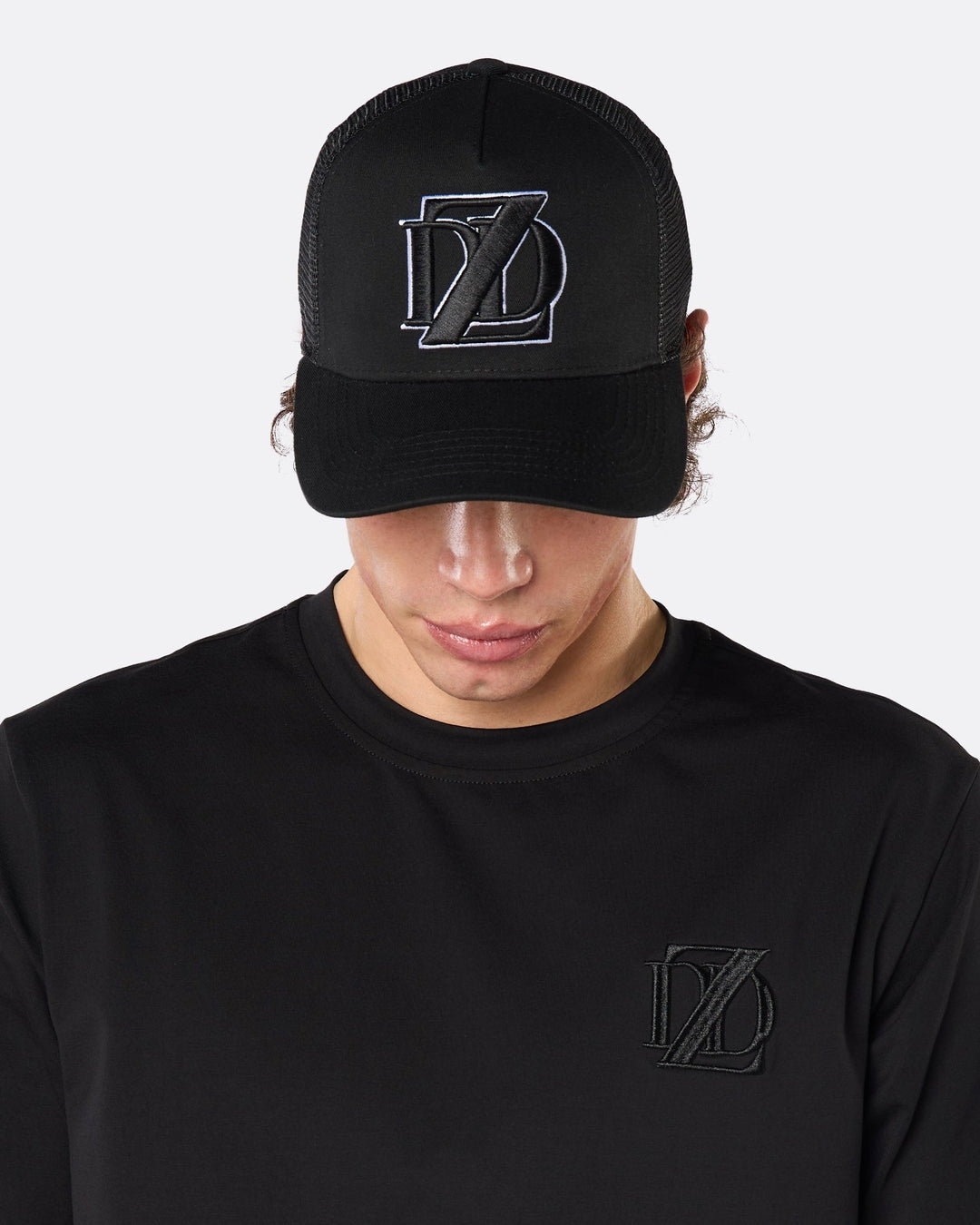 Close-up front view of a male model wearing the DopeDreamz Interlude Noir black trucker cap with tonal black embroidered DDZ monogram, paired with the matching black T-shirt featuring a tonal black embroidered DDZ monogram.