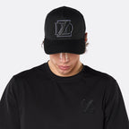 Close-up front view of a male model wearing the DopeDreamz Interlude Noir black trucker cap with tonal black embroidered DDZ monogram, paired with the matching black T-shirt featuring a tonal black embroidered DDZ monogram.