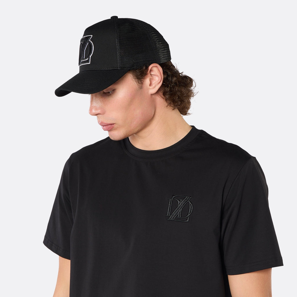 Side profile close-up of a male model wearing the DopeDreamz Interlude Noir black trucker cap with tonal black embroidered DDZ monogram, paired with the matching black T-shirt featuring a tonal black embroidered DDZ monogram.