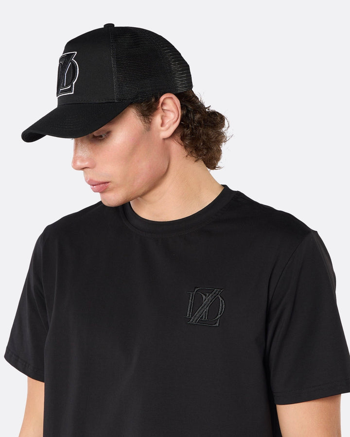 Side profile close-up of a male model wearing the DopeDreamz Interlude Noir black trucker cap with tonal black embroidered DDZ monogram, paired with the matching black T-shirt featuring a tonal black embroidered DDZ monogram.