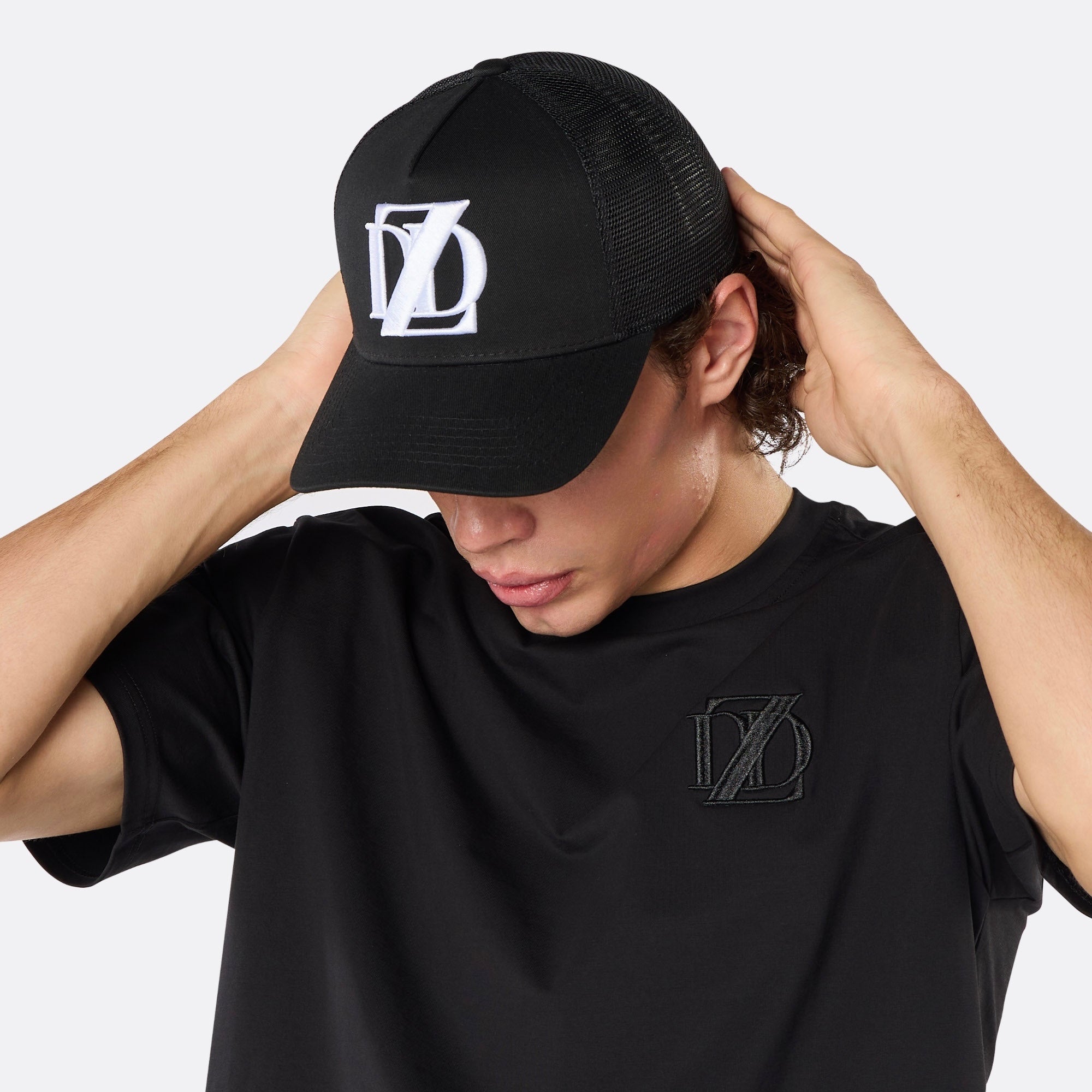 Lifestyle close-up of a male model wearing the DopeDreamz Interlude Noir black trucker cap featuring a premium white embroidered DDZ monogram, paired with the matching black T-shirt featuring a tonal black embroidered DDZ monogram.