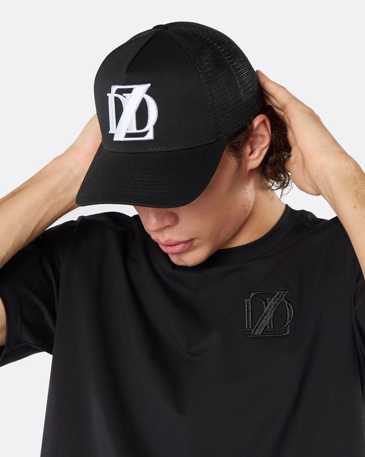 Lifestyle close-up of a male model wearing the DopeDreamz Interlude Noir black trucker cap featuring a premium white embroidered DDZ monogram, paired with the matching black T-shirt featuring a tonal black embroidered DDZ monogram.