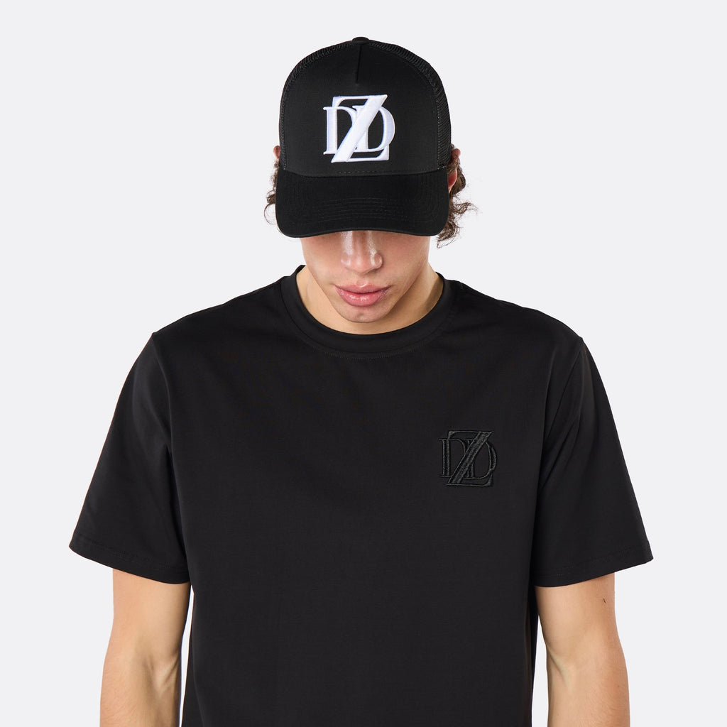 Front close-up of a male model wearing the DopeDreamz Interlude Noir black trucker cap featuring a white embroidered DDZ monogram, paired with the matching black T-shirt featuring a tonal black embroidered DDZ monogram.