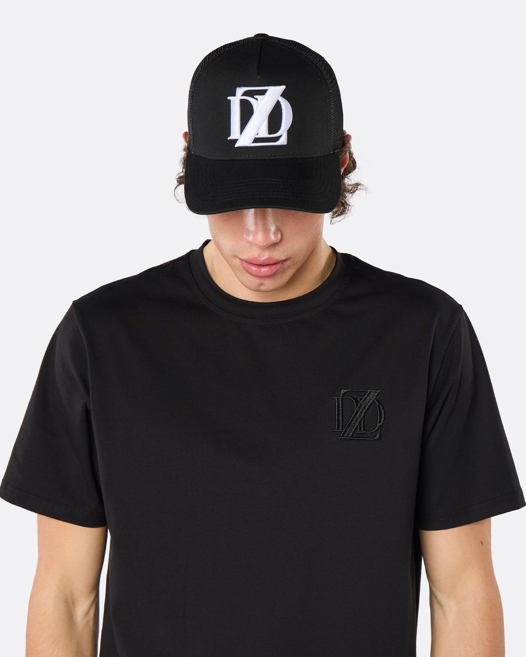 Front close-up of a male model wearing the DopeDreamz Interlude Noir black trucker cap featuring a white embroidered DDZ monogram, paired with the matching black T-shirt featuring a tonal black embroidered DDZ monogram.