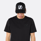 Front close-up of a male model wearing the DopeDreamz Interlude Noir black trucker cap featuring a white embroidered DDZ monogram, paired with the matching black T-shirt featuring a tonal black embroidered DDZ monogram.