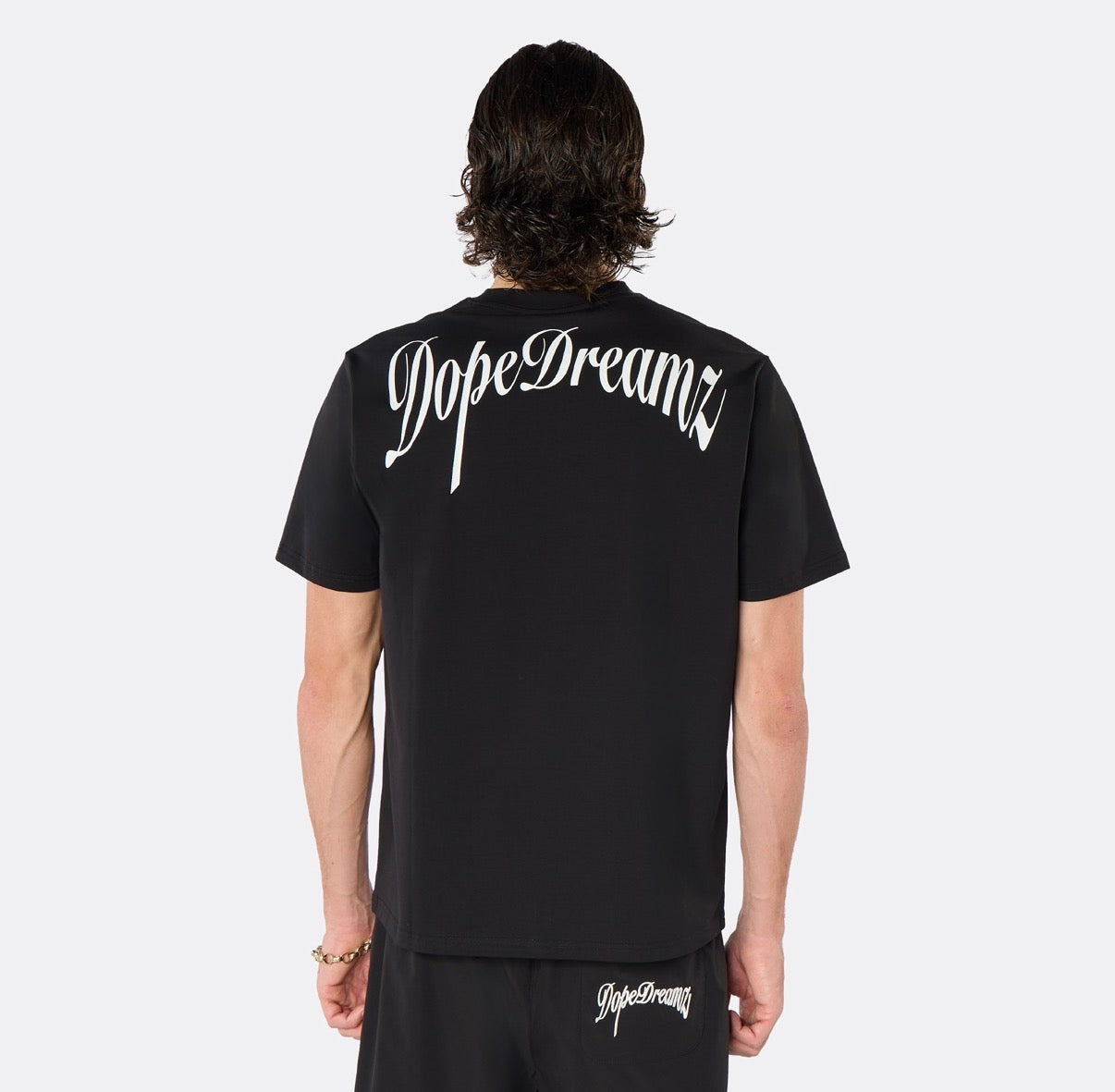 Male model wearing the DopeDreamz Interlude Noir black premium T-shirt with white DopeDreamz back graphic and black shorts with white DopeDreamz logo, back view.