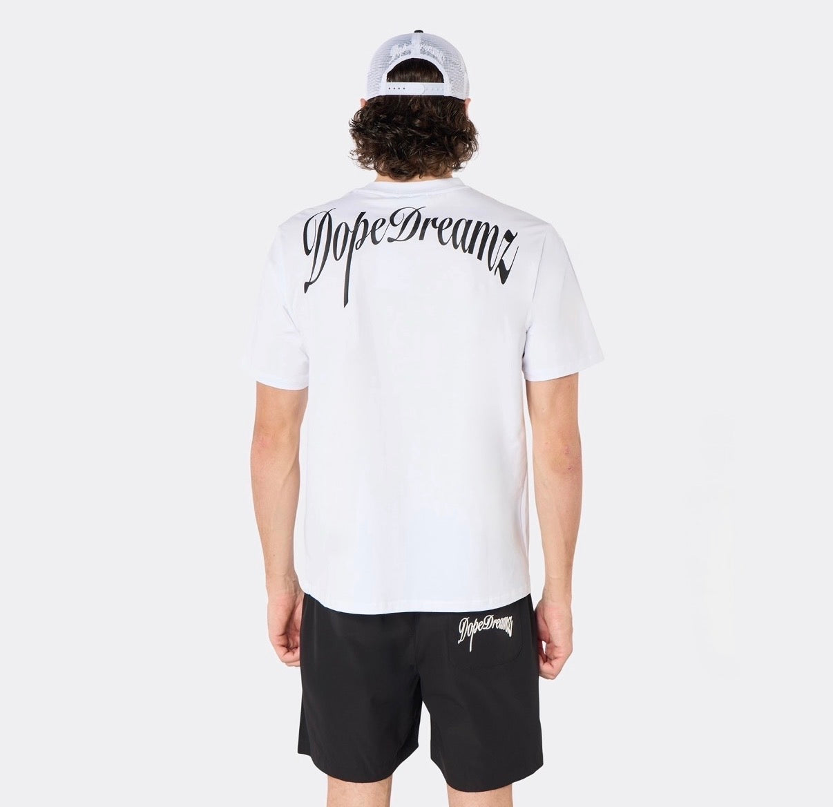 Male model wearing the DopeDreamz Interlude Noir white premium T-shirt with black DopeDreamz back graphic and black shorts with white DopeDreamz logo, back view.